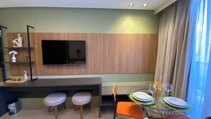 Uma sala de estar moderna com televisão de tela plana fixada em uma parede de painéis de madeira, uma mesa de jantar de vidro com cadeiras e uma pequena área de estar com dois banquinhos acolchoados.