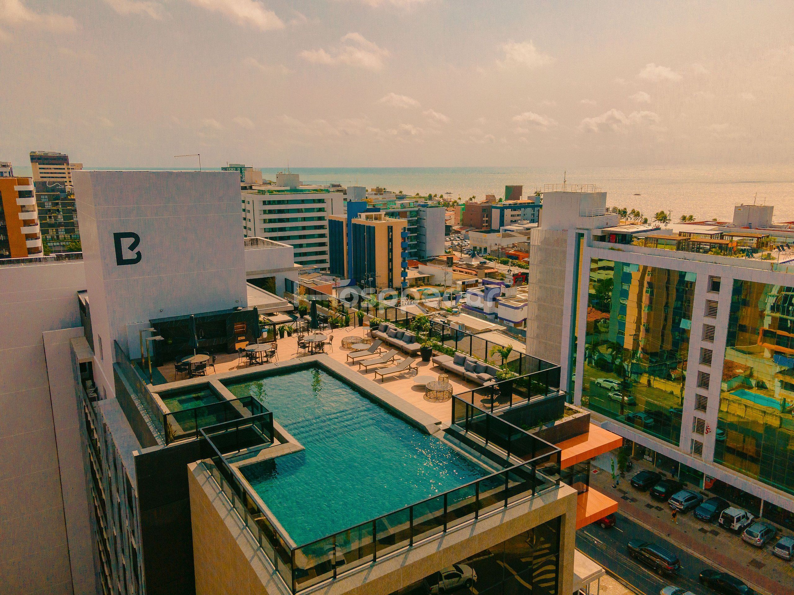 A rooftop pool with a glass barrier, surrounded by lounge chairs and umbrellas, overlooking a cityscape with buildings and a body of water.