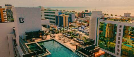 A rooftop pool with a glass barrier, surrounded by lounge chairs and umbrellas, overlooking a cityscape with buildings and a body of water.