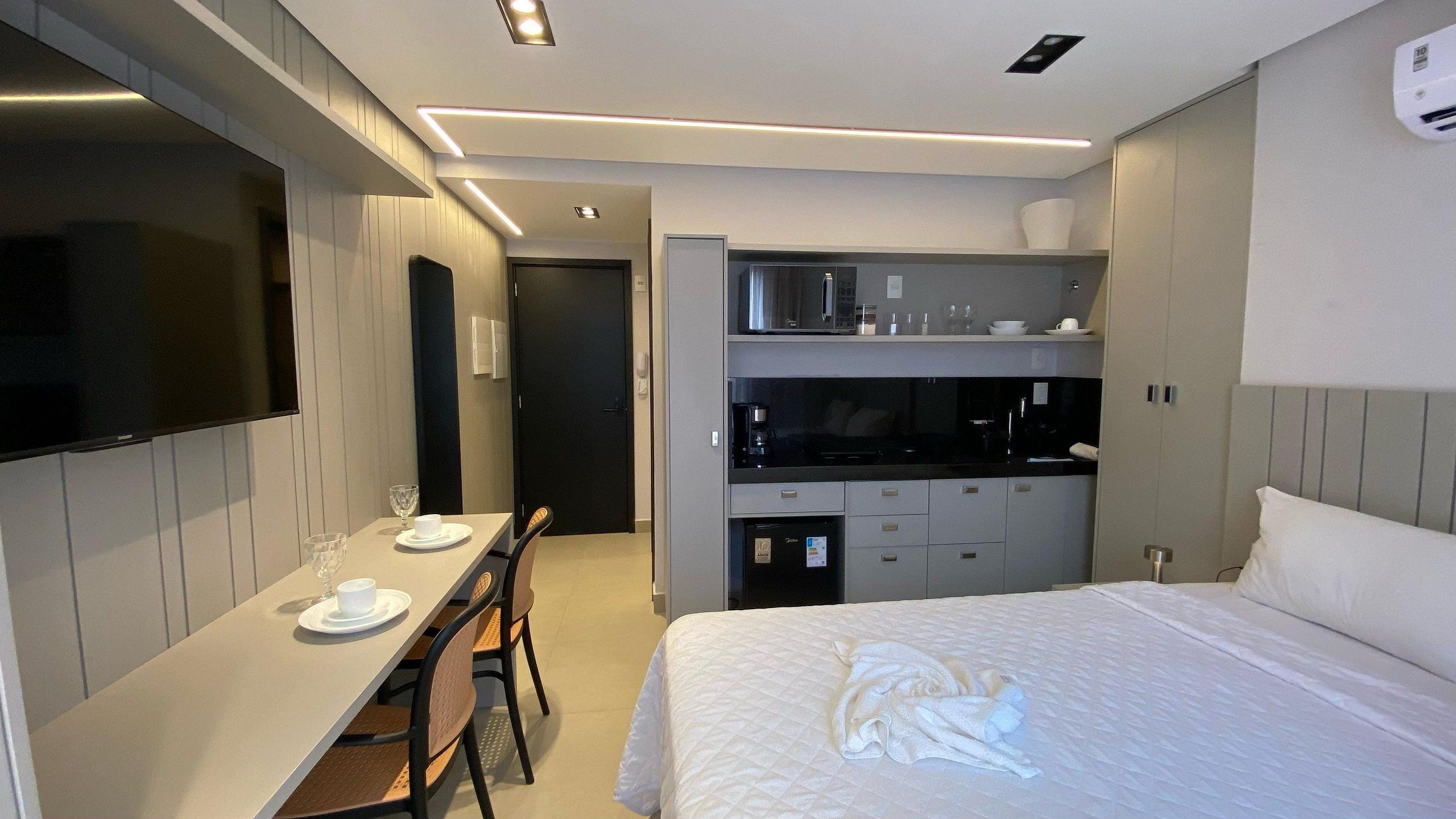 Quarto de hotel com cama, mesa de jantar com cadeiras, cozinha compacta e televisão.