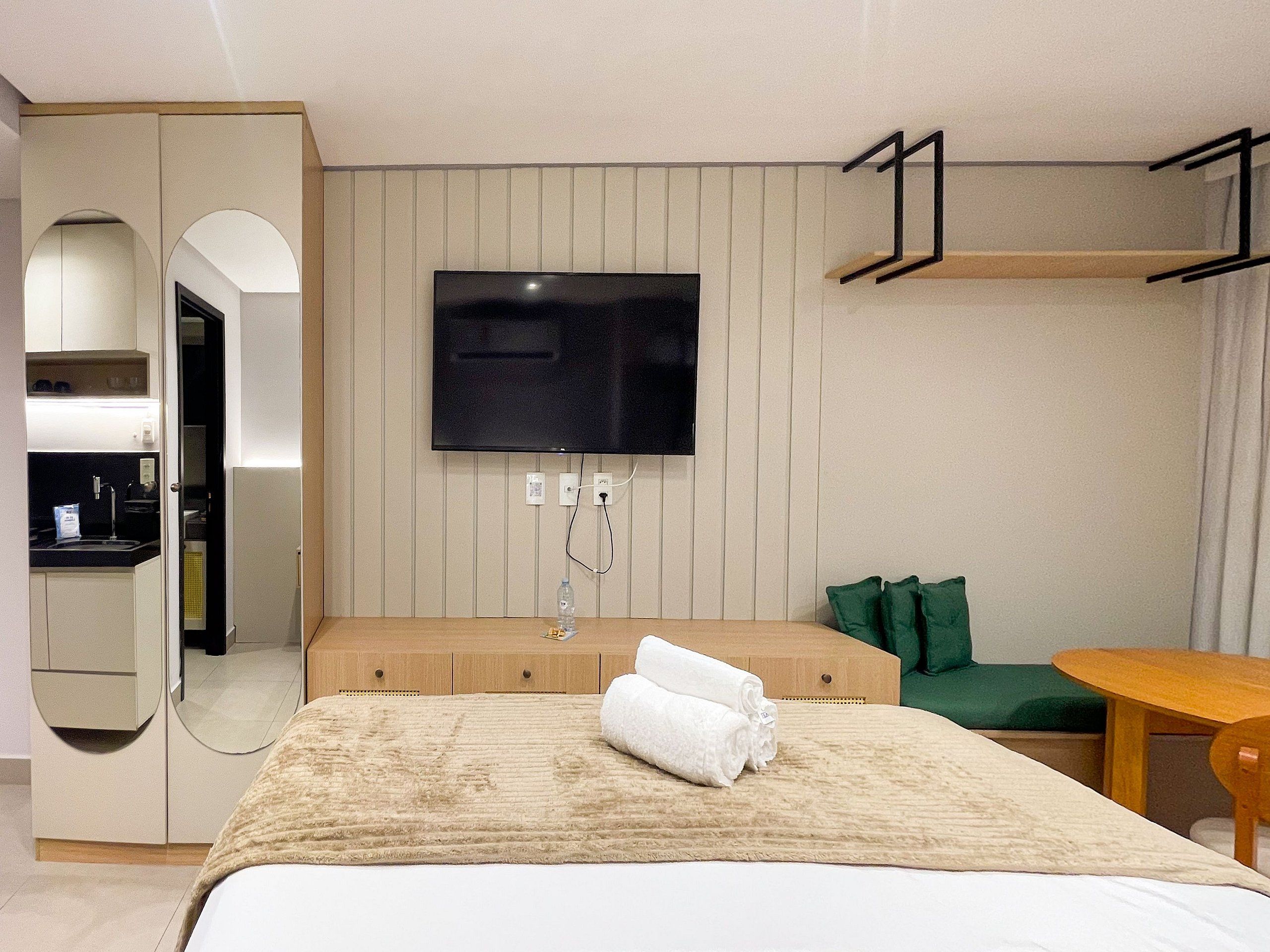 Quarto de hotel com uma cama, televisão presa na parede, pequeno espaço de cozinha e uma cadeira verde.