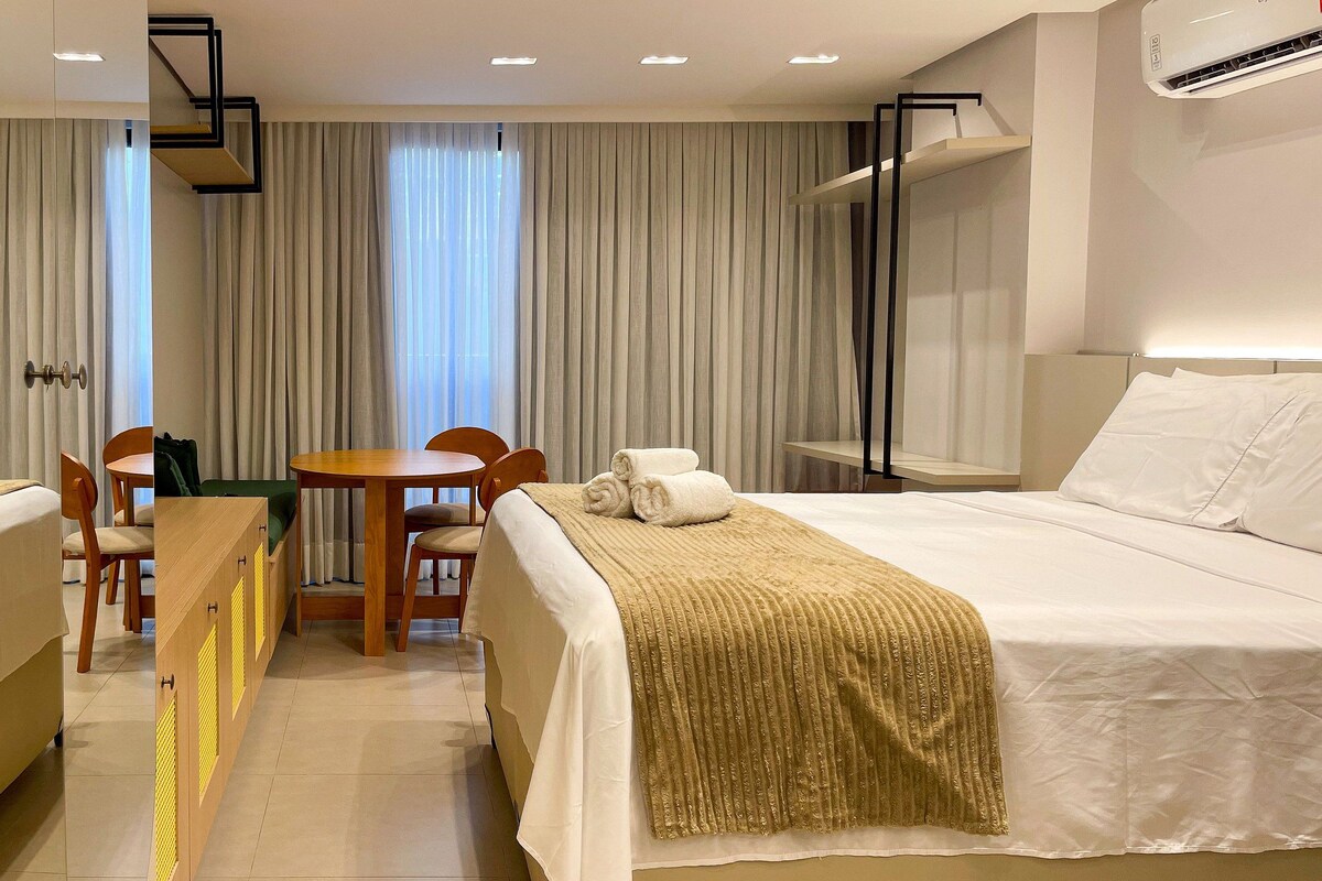 Quarto de hotel com cama, escrivaninha, cadeiras, janela com cortinas, ar condicionado e prateleira na parede.
