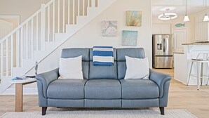 Ein modernes Wohnzimmer mit einem grauen Sofa, weißen Kissen und einer gestreiften Decke. Eine Treppe mit weißen Geländern führt zu einem Küchenbereich mit Edelstahlgeräten.
