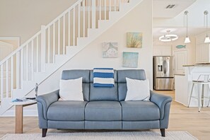 Ein modernes Wohnzimmer mit einem grauen Sofa, weißen Kissen und einer gestreiften Decke. Eine Treppe mit weißen Geländern führt zu einem Küchenbereich mit Edelstahlgeräten.