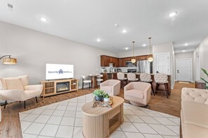 Ein modernes Wohnzimmer mit Kamin, einem Essbereich und einer dahinterliegenden Küche, sowie einem Fernseher, der ein Logo zeigt.