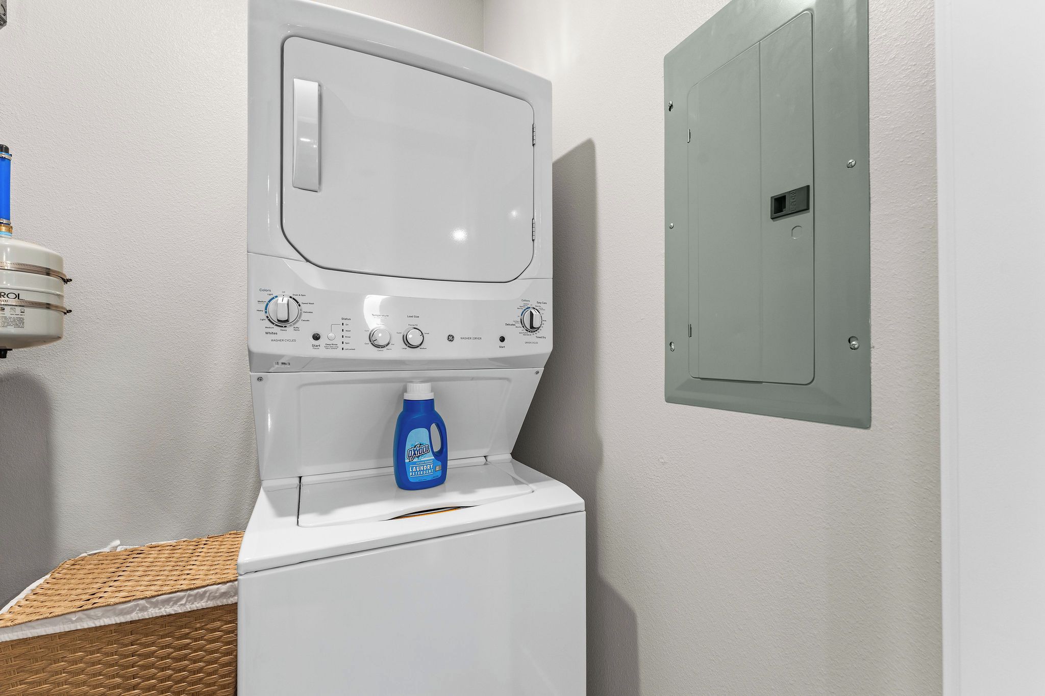 Een voorlader wasmachine met een fles wasmiddel er bovenop, een bedieningspaneel met diverse instellingen, en een gas- en watermeter aan de muur.