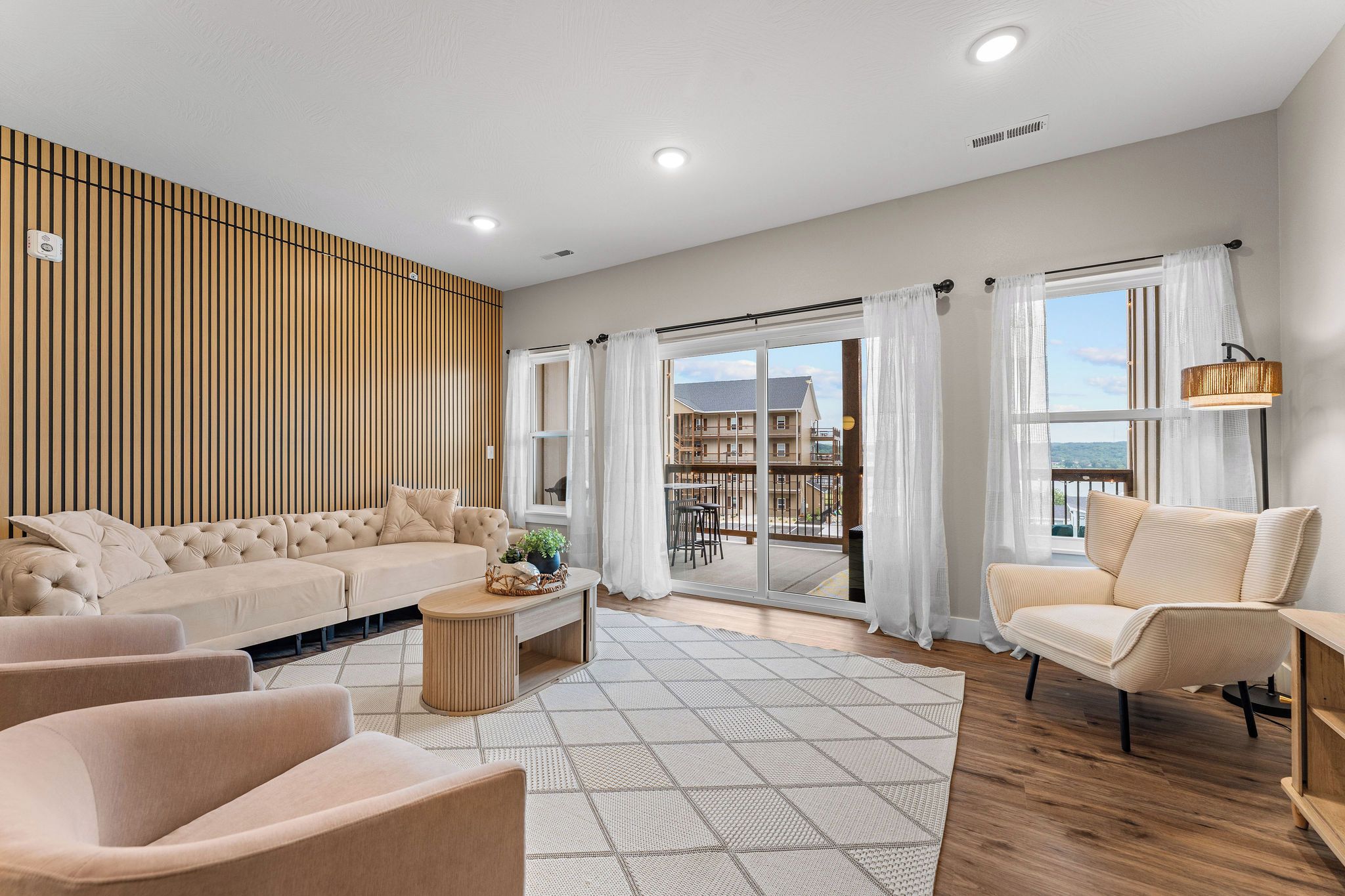 A modern living room with a sectional sofa, armchairs, a coffee table, and a view of a balcony with outdoor furniture.