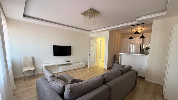 A modern living room with a grey sectional sofa, a flat-screen TV, a white entertainment unit, and a kitchen area in the background. - Bright Modern 2BR Apt near Central Park & Beach (Batumi)