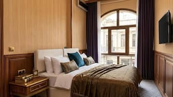 Quarto de hotel com uma cama grande, mesa de cabeceira com livros, televisão e vista para um edifício pela janela.