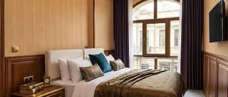 Habitación de hotel con una cama grande, una mesita de noche con libros, un televisor y vistas a un edificio desde la ventana.