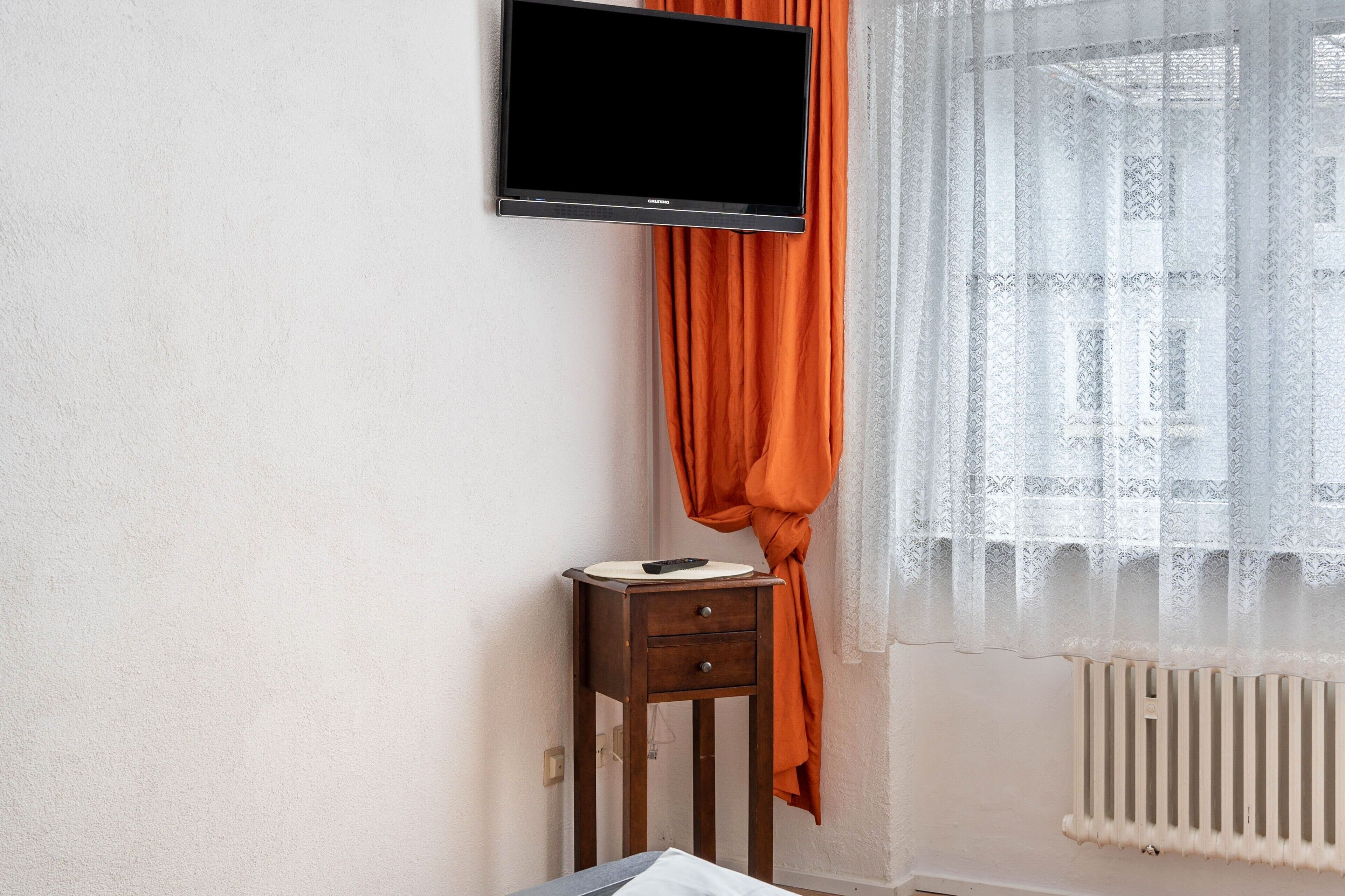 Ein Zimmer mit an der Wand befestigtem Fernseher, einem kleinen Nachttisch mit Fernbedienung und einem Fenster mit durchsichtigen Vorhängen.