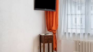 Ein Zimmer mit an der Wand befestigtem Fernseher, einem kleinen Nachttisch mit Fernbedienung und einem Fenster mit durchsichtigen Vorhängen.