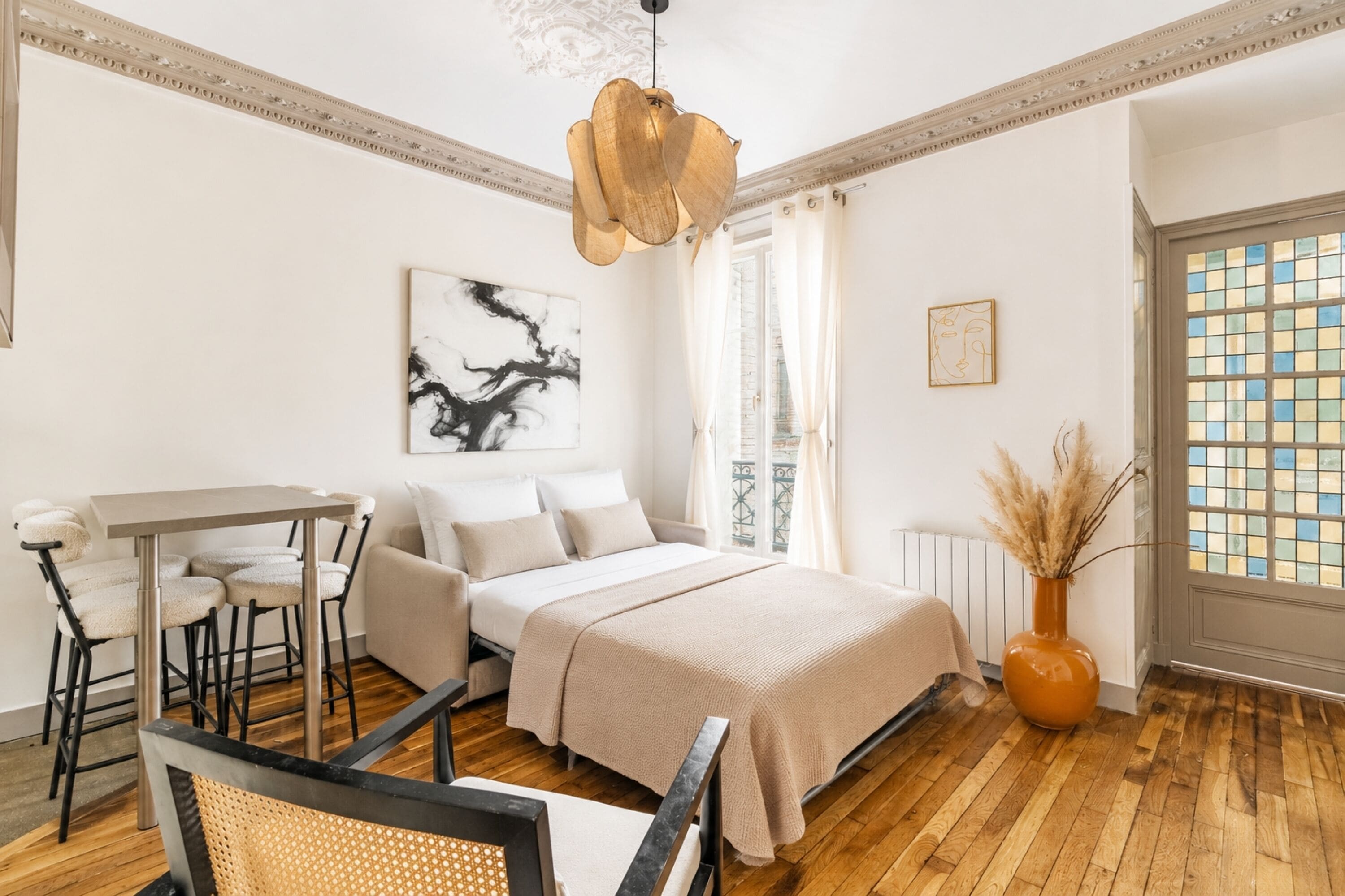 Ein Schlafzimmer mit Bett, Schreibtisch und Stuhl, ein Fenster mit Blick und eine Vase mit getrockneten Pflanzen.