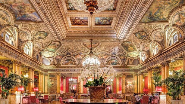 A grand lobby with ornate ceiling frescoes, a central wooden table, and lush potted plants.