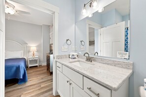 A modern bathroom with a large mirror, a sink, and a toilet. There is a bed with a blue blanket in the adjacent room. - Circle Pearl-Ocean View Townhome (Atlantic Beach)