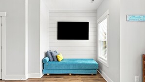 A room with a blue sofa, a flat-screen TV mounted on the wall, and a window with blinds. - Circle Pearl-Ocean View Townhome (Atlantic Beach)