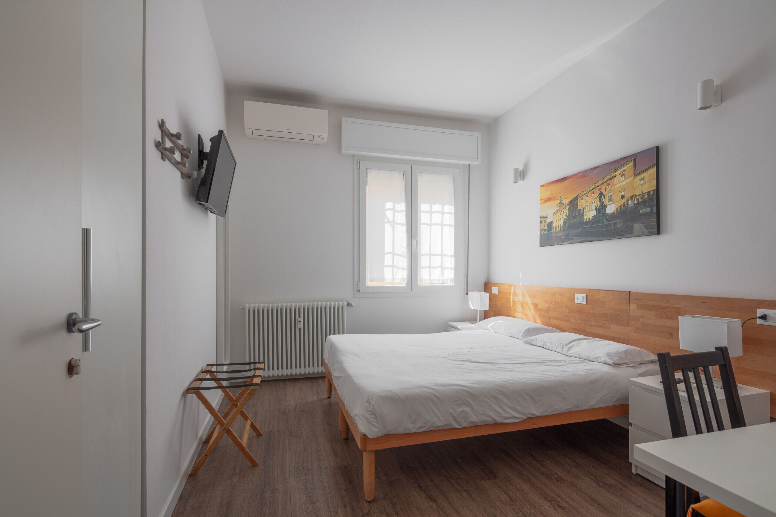 Een slaapkamer met een bed, bureau, stoel, radiator, raam en een schilderij aan de muur.