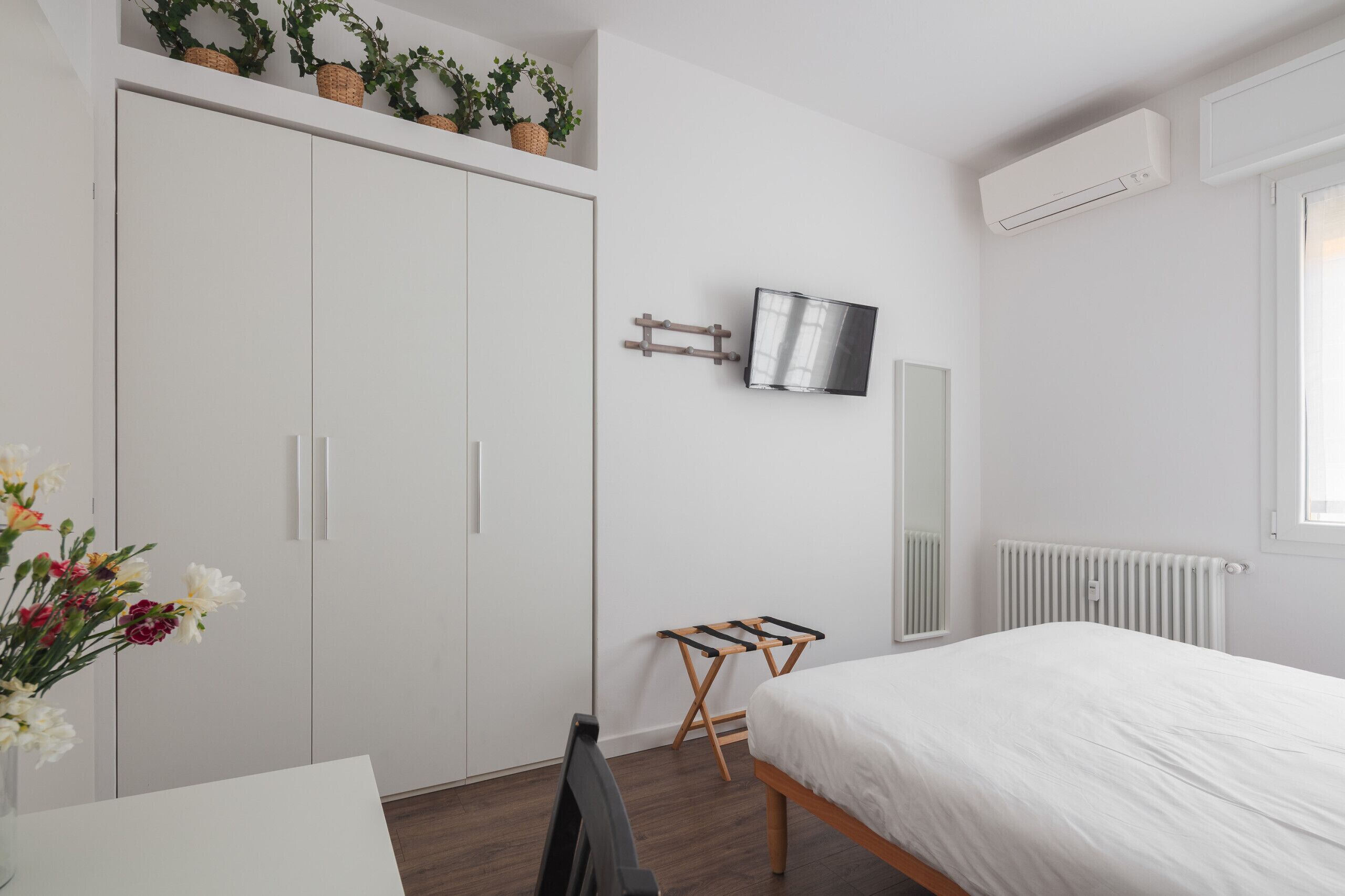 Een slaapkamer met een bed, een televisie, een radiator, een kledingkast en een kleine tafel met een stoel.
