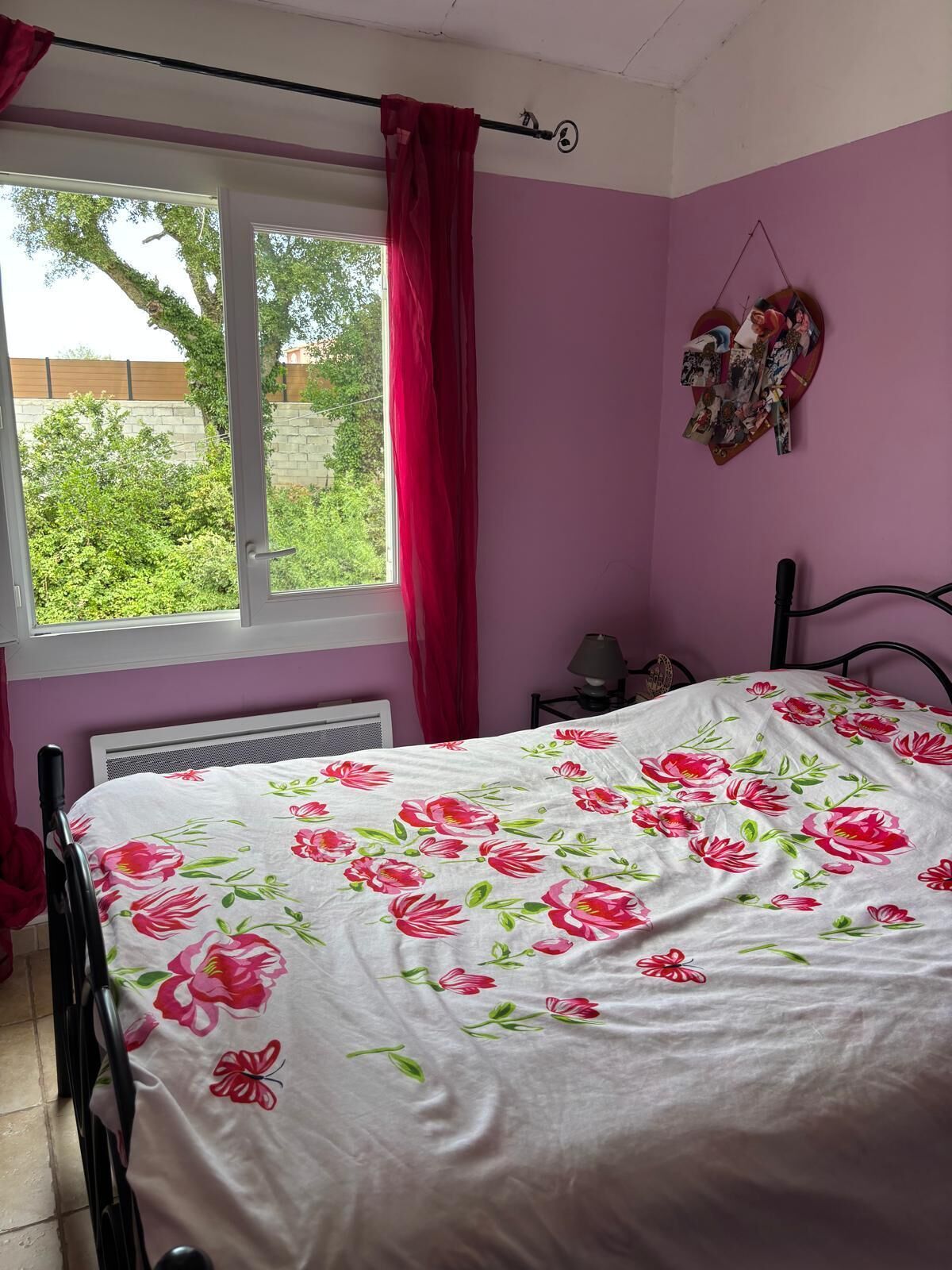 A bedroom with a bed, a window with curtains, a radiator, and a wall-mounted heart decoration.