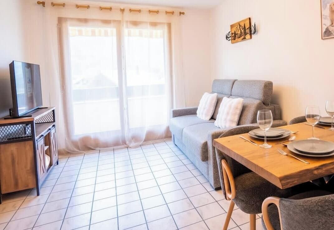 Een woonkamer met een bank, een eettafel gedekt voor twee personen, een televisie op een standaard en een raam met gordijnen.