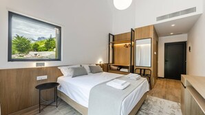 A modern bedroom with a large bed, a small stool, a wardrobe, and a view of greenery outside.