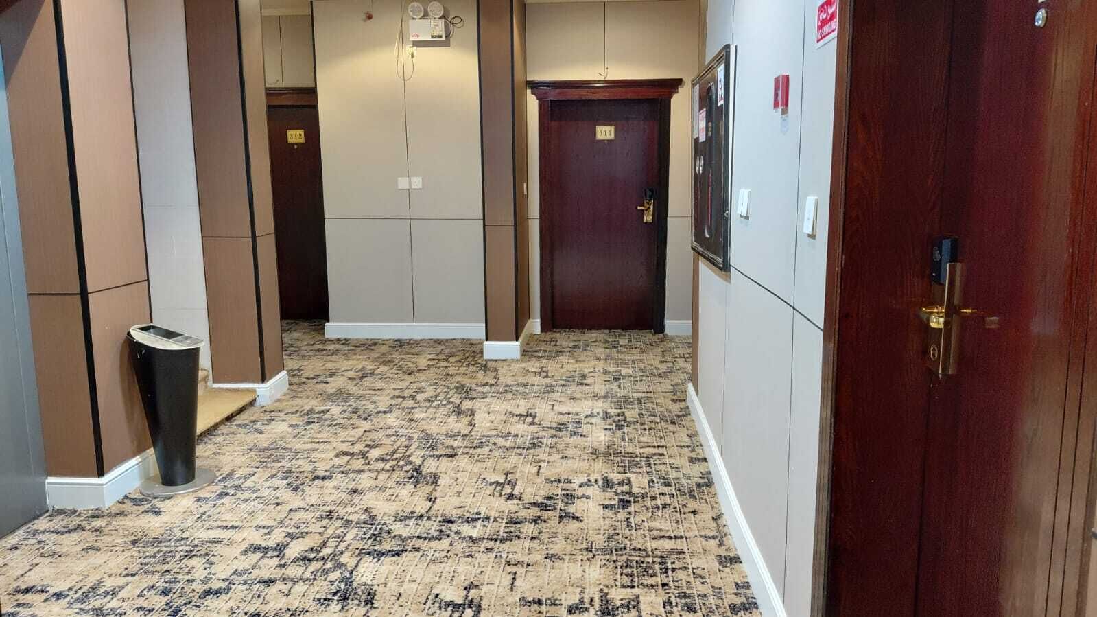 A hallway with a patterned carpet, a trash bin, a wooden door, and a wall-mounted light fixture.