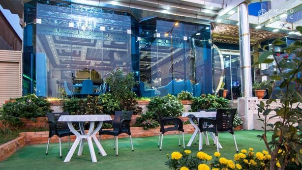 An outdoor seating area with tables and chairs, surrounded by greenery and yellow flowers.