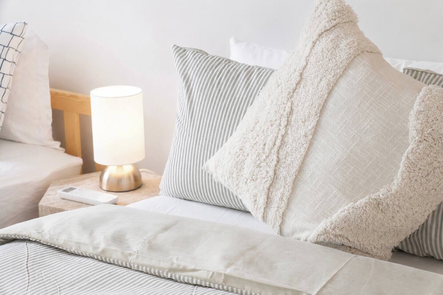 Ein ordentlich bezogenes Bett mit einem weiß-grauen Kissen, einer weißen Decke und einer Nachttischlampe auf einem Holznachttisch.