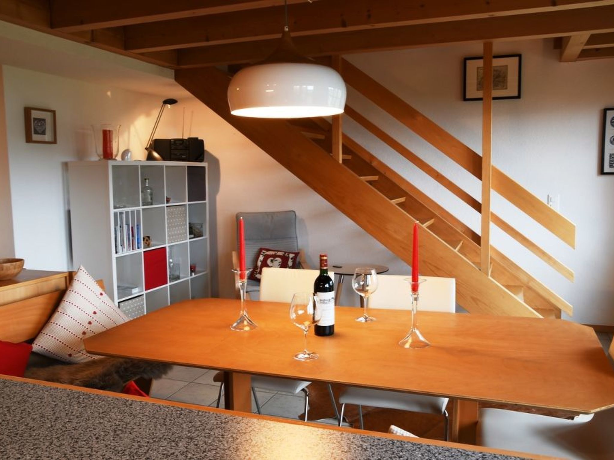 Un comedor con una mesa de madera, copas de vino y una botella. Se ve una escalera al fondo.