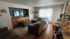 A living room with a TV, sofa, ottoman, and a kitchen area with bar stools. - Spacious 5-bedroom house in Cheyenne with AC, fitness room, outdoor space (Cheyenne)