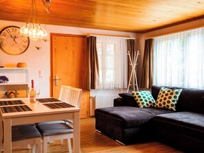 Ein gemütliches Wohnzimmer mit Holzboden, einem Esstisch mit Stühlen, einer Couch mit gemusterten Kissen und einer großen Wanduhr.