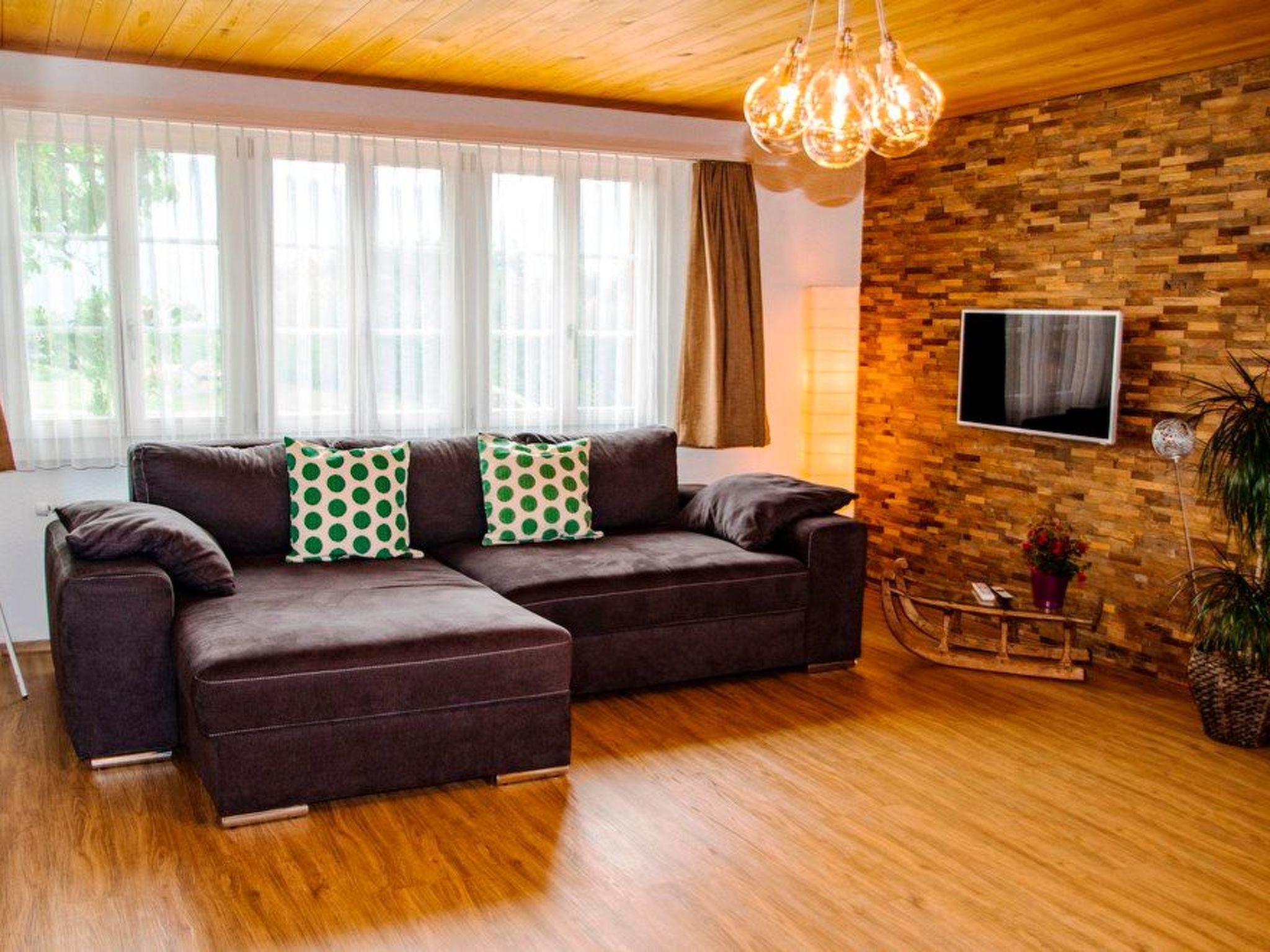 A modern living room with a sectional sofa, wooden flooring, a stone wall, a TV mounted on the wall, and a hanging light fixture.
