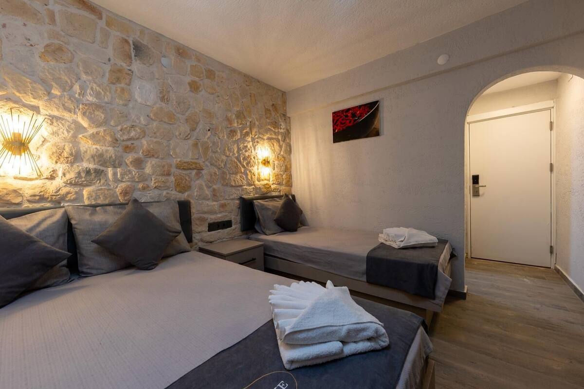 Una habitación con pared de piedra, dos camas con almohadas, una lámpara fijada a la pared, una puerta y una toalla sobre la cama.
