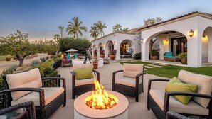 Un patio exterior con mobiliario de mimbre, un hogar central y un edificio con puertas arqueadas y asientos al aire libre.