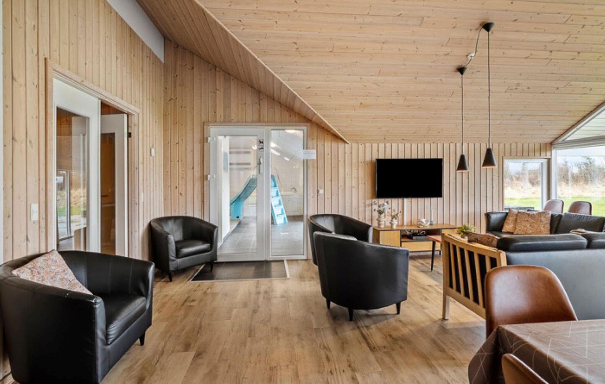 Een ruime woonkamer met houten wanden, een schuifdeur naar een balkon, een flatscreen televisie aan de muur en een eethoek met een tafel en stoelen.