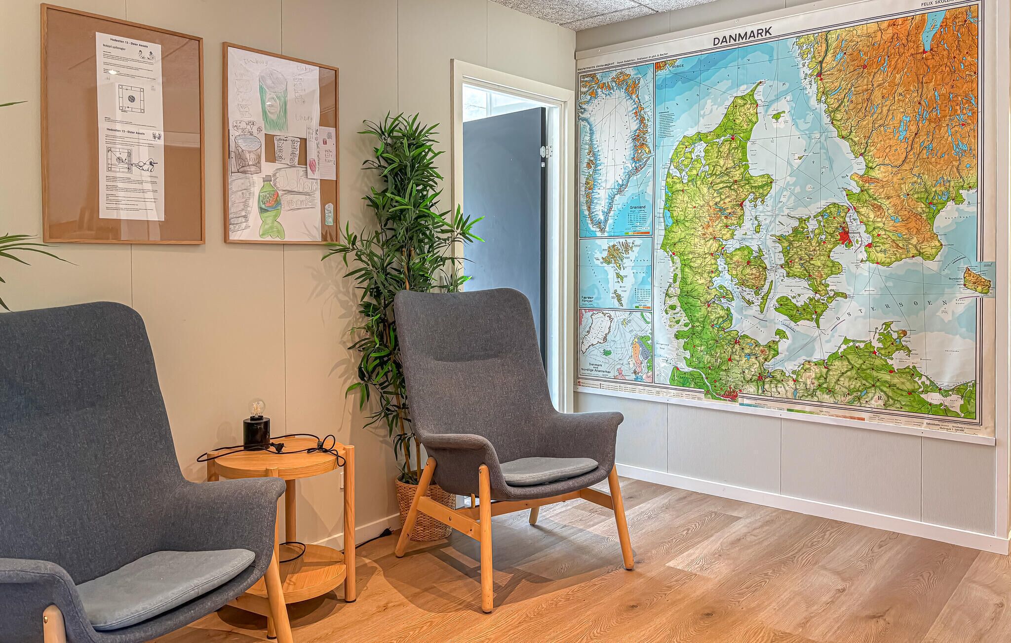 A room with two grey armchairs, a wooden side table, a large map of Denmark, and a bulletin board with papers.