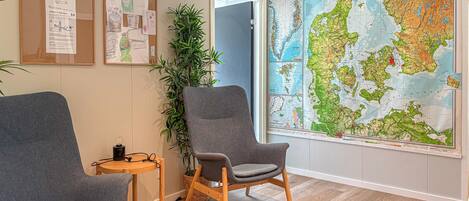 A room with two grey armchairs, a wooden side table, a large map of Denmark, and a bulletin board with papers.