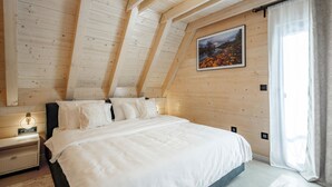 Een slaapkamer met een schuine plafond, een bed met witte beddengoed, een nachtkastje met een lamp en een ingelijst schilderij aan de muur.