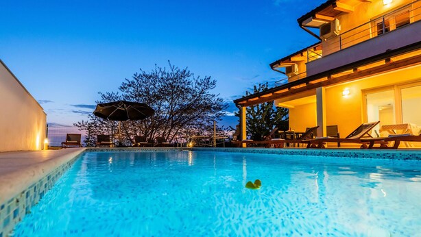Una piscina con sillas de descanso y sombrillas, un edificio con balcones y un árbol al fondo.