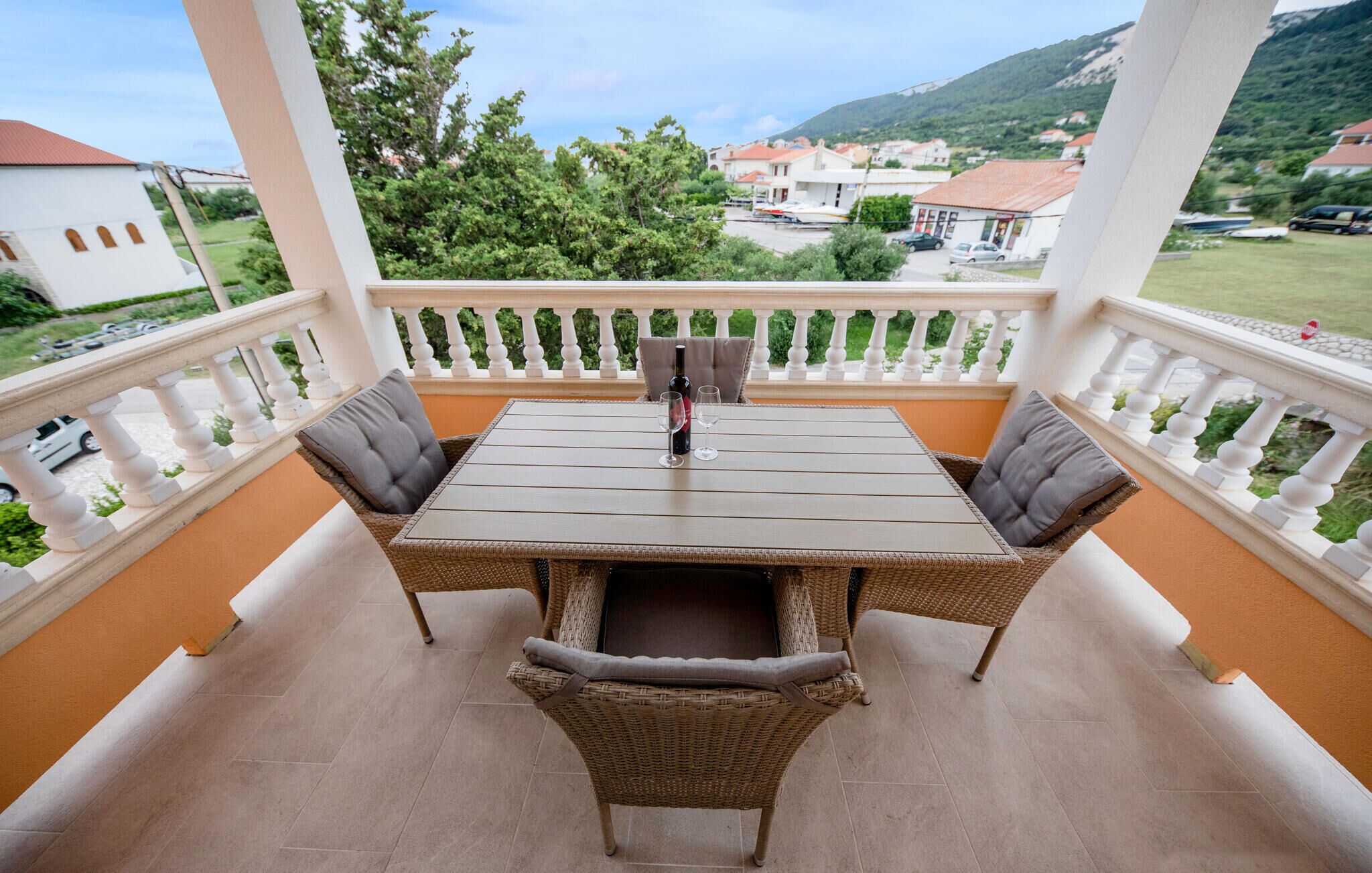 A balcony with a table and chairs, a bottle of wine, and a view of a town and mountains.
