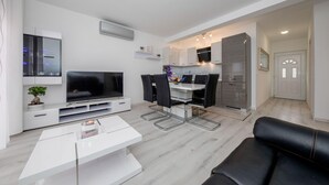 Ett modernt vardagsrum med en platt-TV, ett matbord med stolar, en svart lädersoffa och en köksdel med skåp och ett kylskåp.