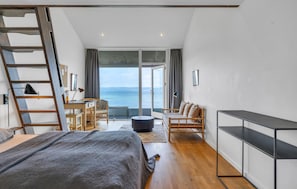 Quarto com cama, escrivaninha, poltrona, sofá, rack para TV e vista para o mar.