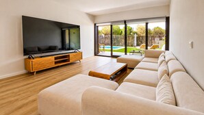 En moderne stue med et stort fladskærms-tv, en træunderholdningsenhed, beige sofaer og en skydedør til en pool.