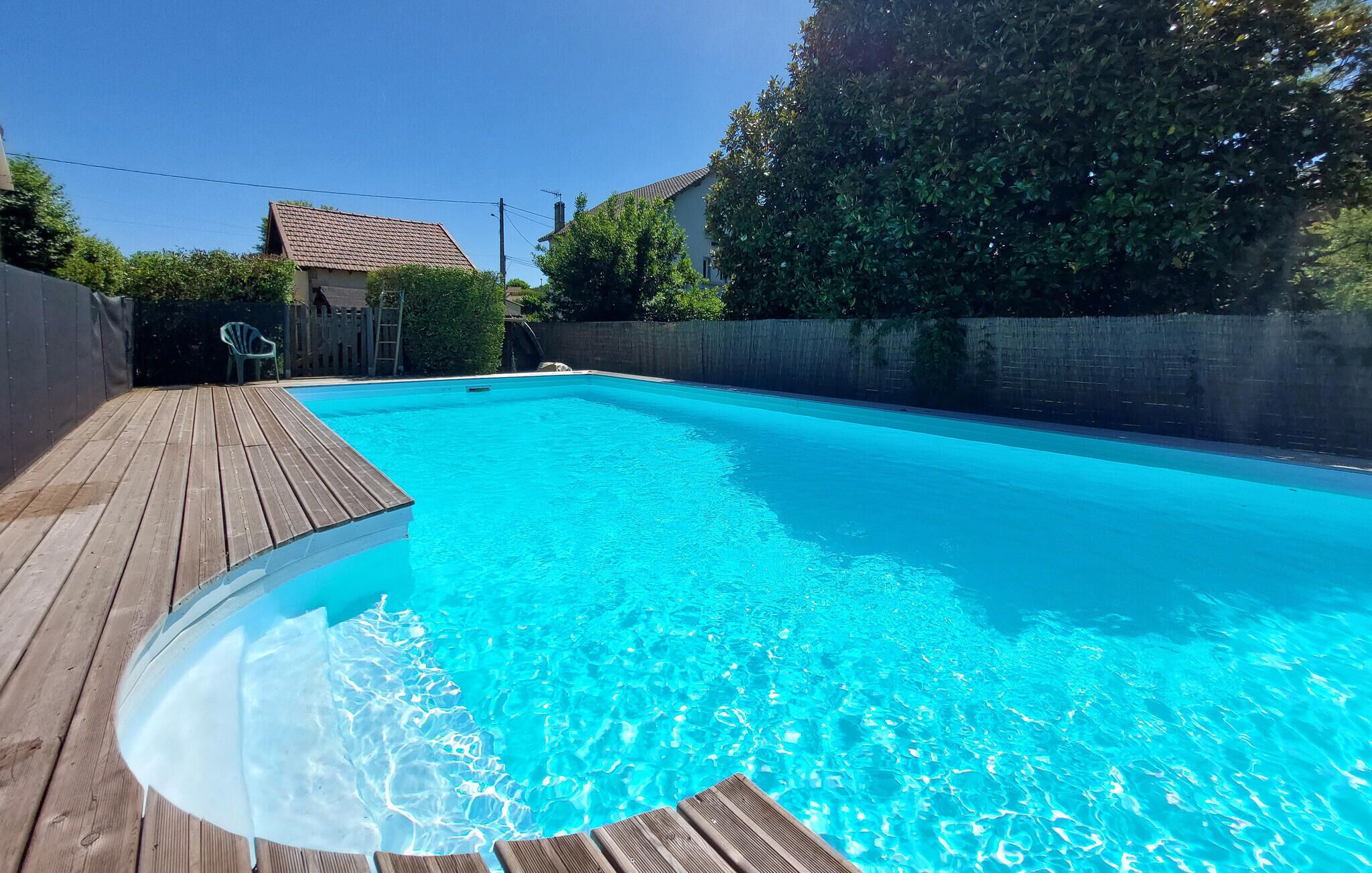 A rectangular swimming pool with a wooden deck, surrounded by a low stone wall and trees.