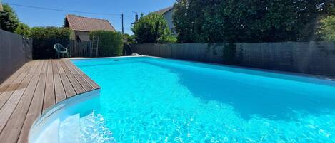 A rectangular swimming pool with a wooden deck, surrounded by a low stone wall and trees.