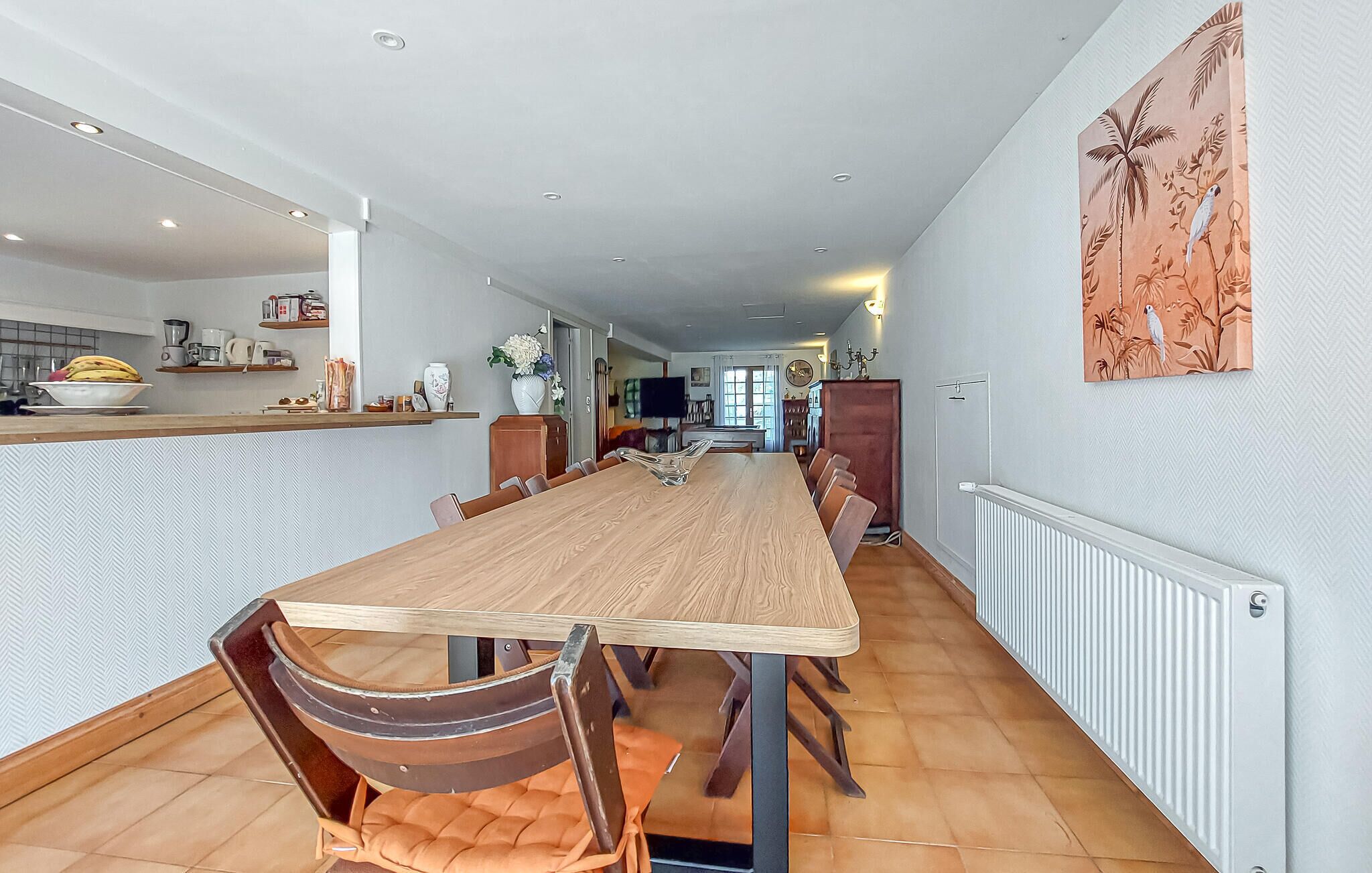Ein großzügiger Essbereich mit einem langen Holztisch und mehreren Stühlen, eine Küchenzeile mit Arbeitsfläche und verschiedenen Geräten sowie ein Wohnzimmer mit Fernseher und Sitzgelegenheiten.