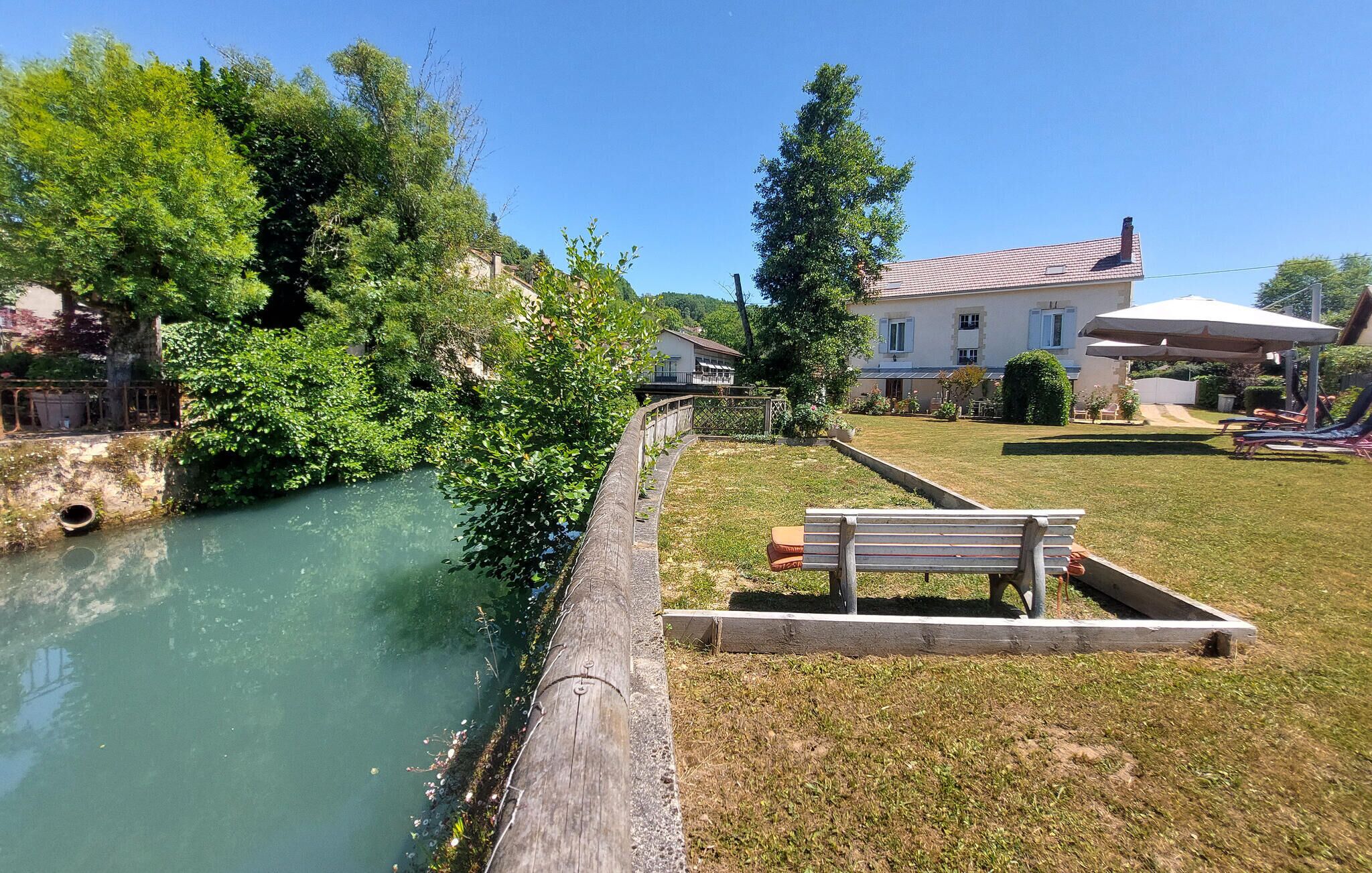 A riverside property with a grassy lawn, a wooden bench, and a house with a tiled roof.