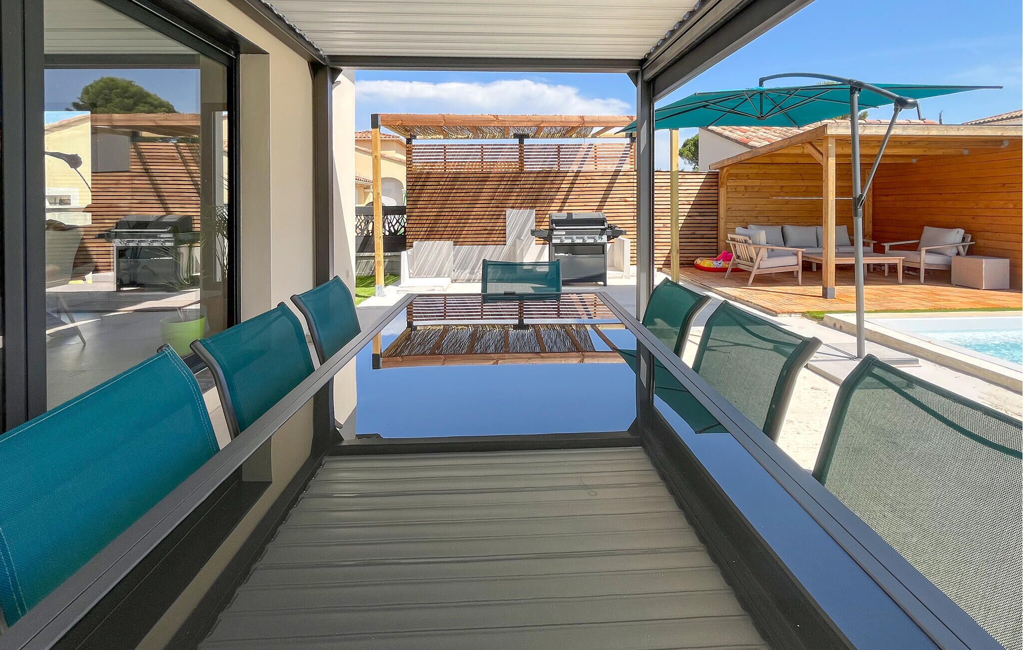 Un espace extérieur aménagé, abrité et doté d’une table et de chaises, d’un barbecue et d’une piscine en arrière-plan.