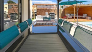 Ein überdachter Außenbereich mit Sitzgelegenheiten, einem Tisch, Stühlen, einem Grill und einem Pool im Hintergrund.