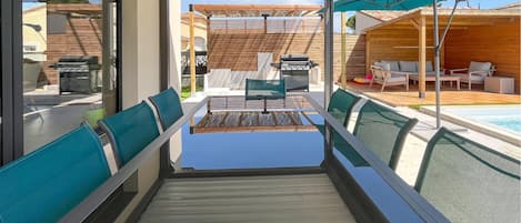 Un espacio de estar techado al aire libre con mesa y sillas, una parrilla y una piscina al fondo.